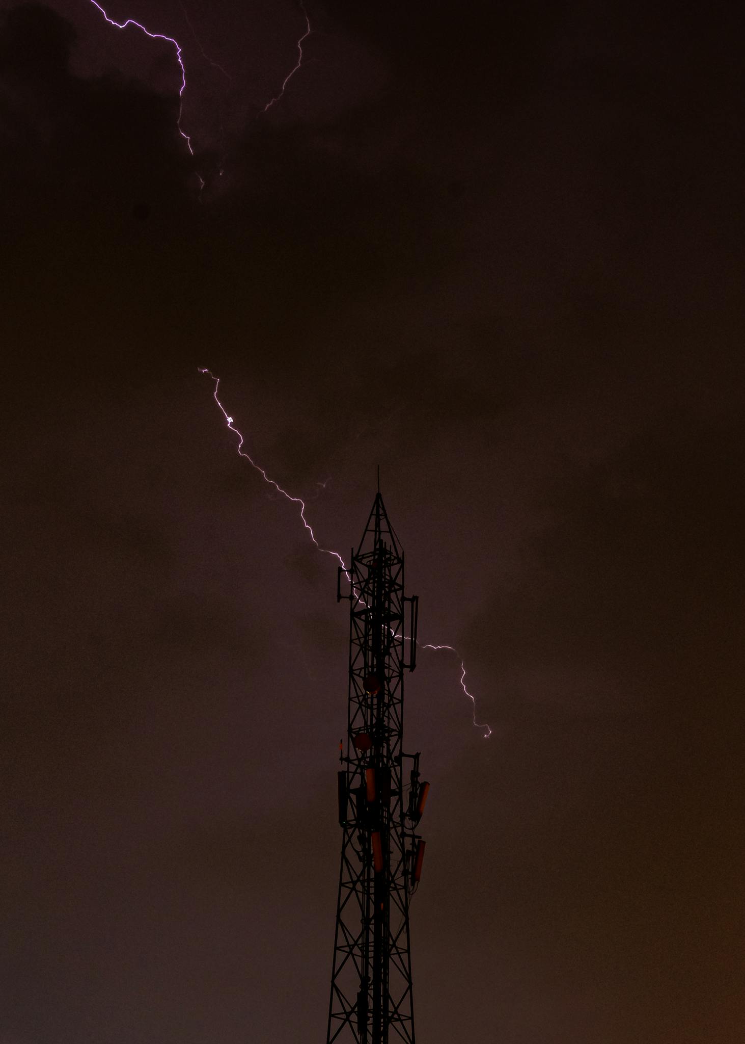 Thunderstorm with lightning striking near a silhouetted transmission tower at night.